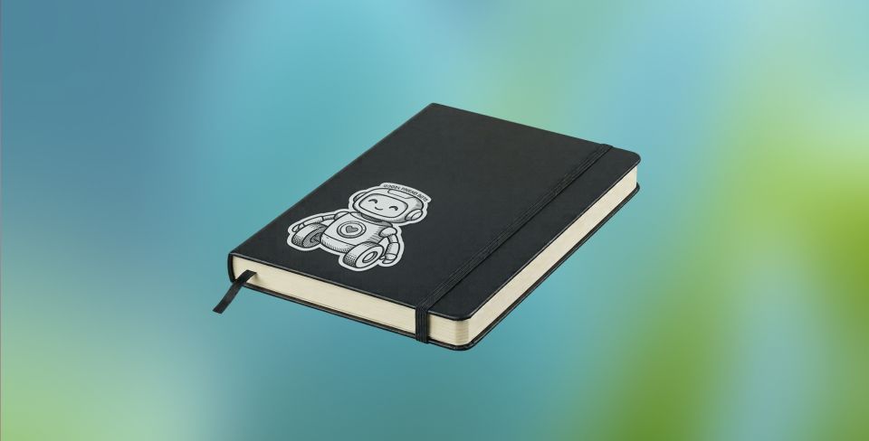 A black hardcover notebook with an elastic band and a sticker of a smiling robot on the cover, against a blurred blue and green background.